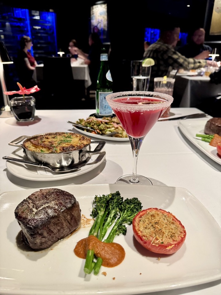A steakhouse plate with a filet mignon, broccolini, and a baked stuffed tomato, with a red cocktail and a dish of potatoes in the background.